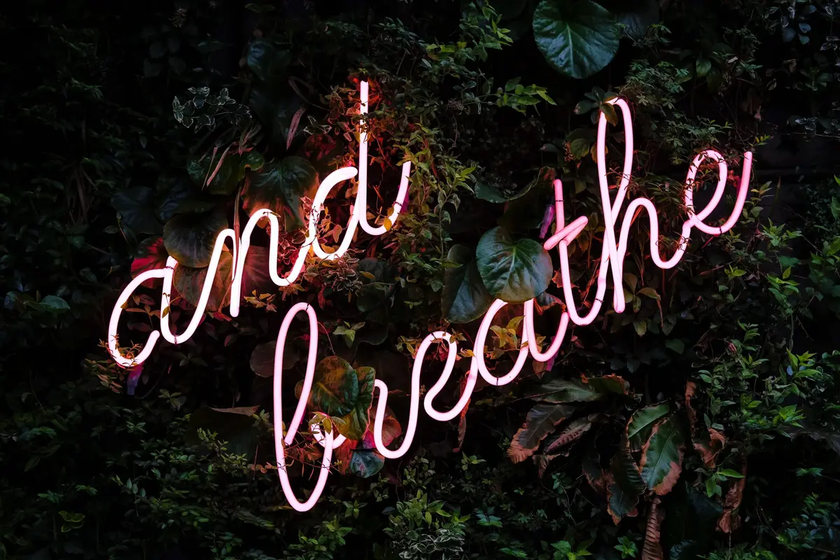 Pink neon sign reading “and breathe” set against a wall of dark green leaves
